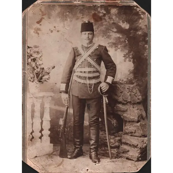 Bir elinde kılıcı diğer elinde tüfek ve fişekleriyle poz veren kalpaklı genç Osmanlı askerinin kabin fotoğrafı, 11x14 cm